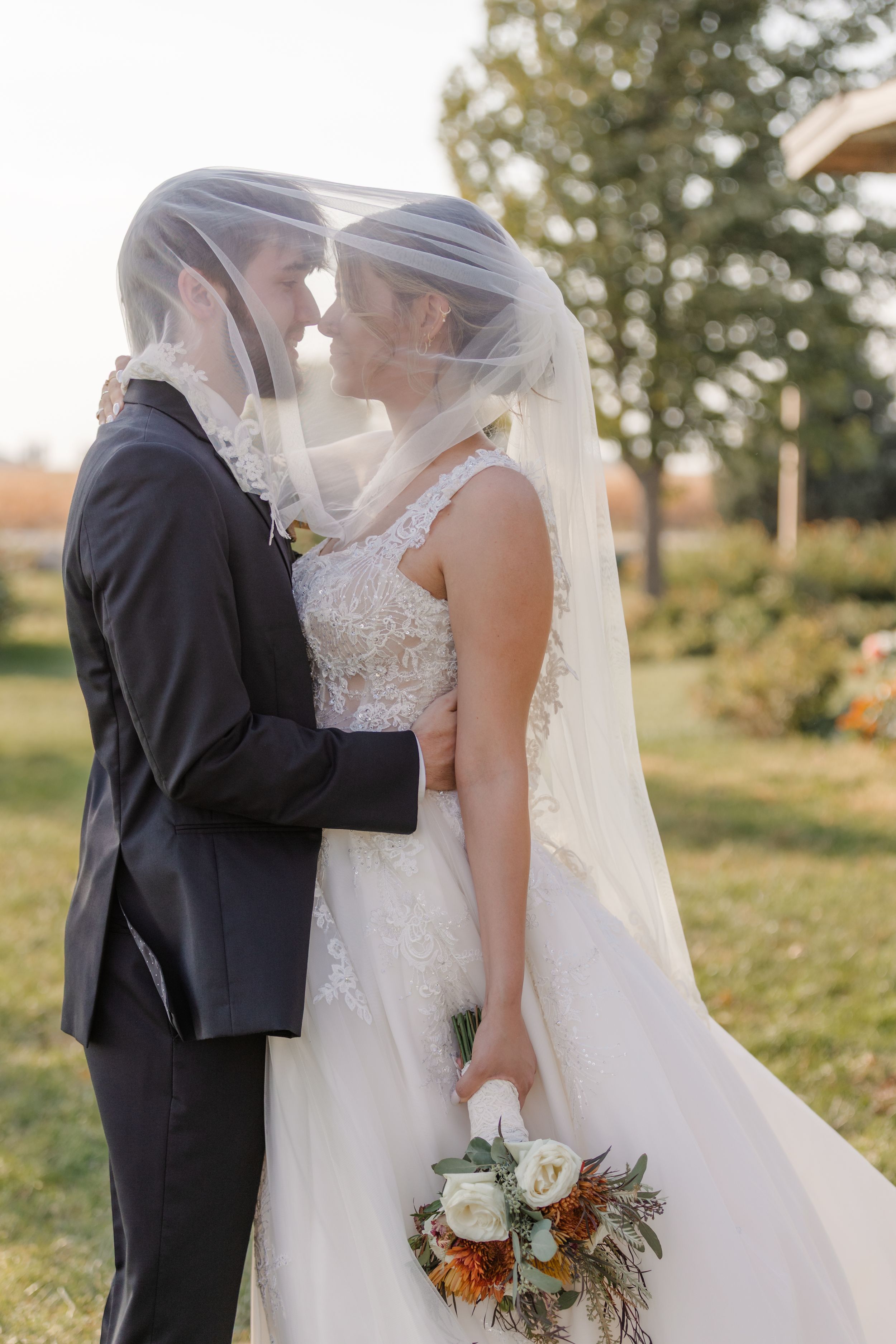 A bride and groom share an intimate kiss outdoors while holding a rustic autumn bouquet with flowing veil.
