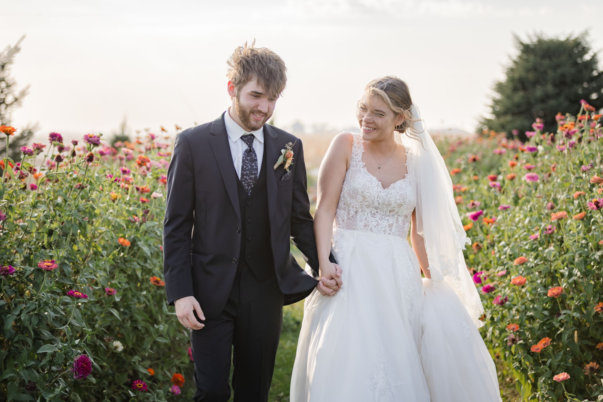 A couple walks hand in hand through a vibrant field of red and pink wildflowers on their wedding day.