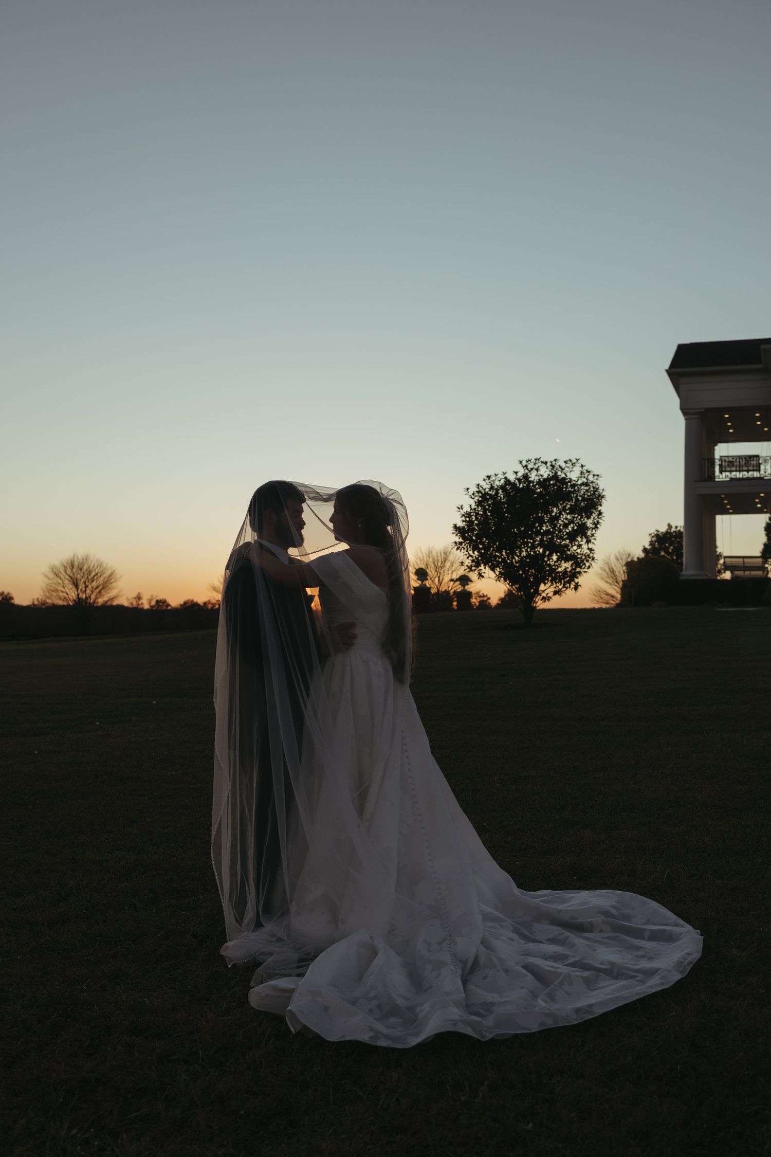A couple in wedding attire share an intimate silhouette moment against a sunset sky in a field.