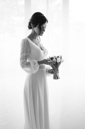 Elegant silhouette of a bride in a flowing white dress holding a bouquet against a bright window.