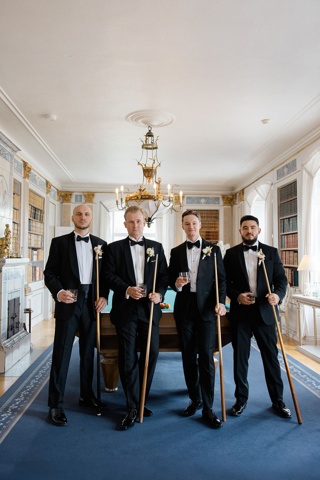 Groomsmen in black tuxedos holding drinks pose together on blue carpet in elegant hallway with bookshelves.