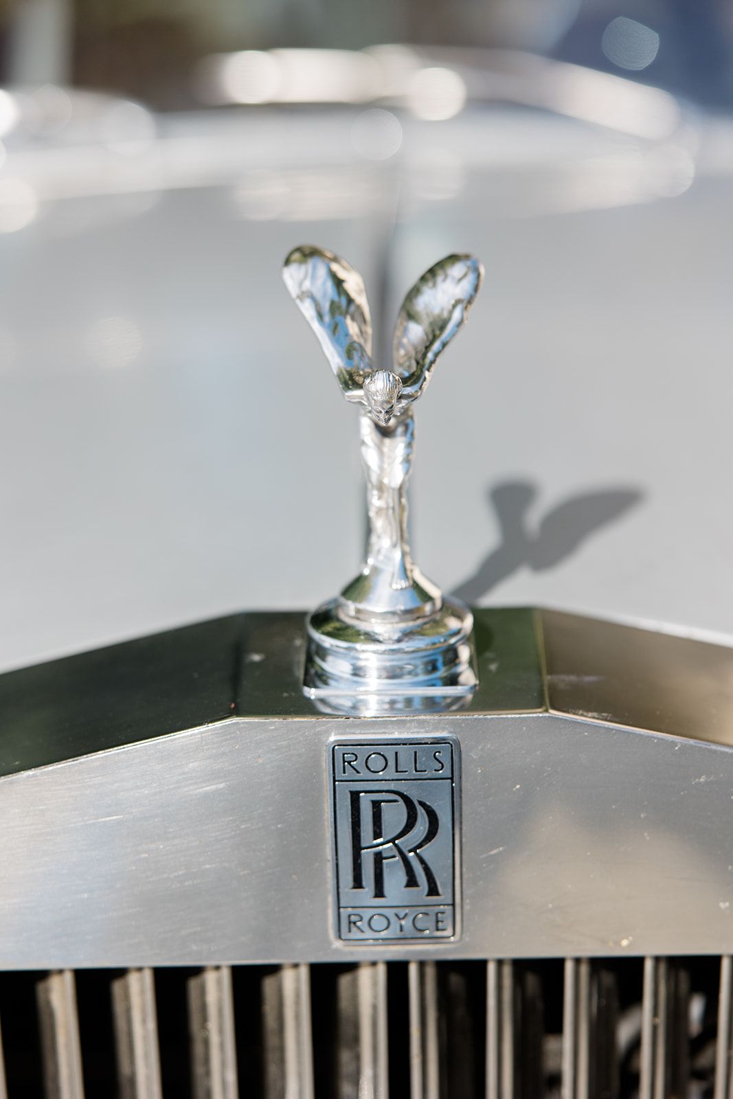 Close-up of iconic Rolls-Royce Spirit of Ecstasy hood ornament and RR logo on silver grille in soft focus.