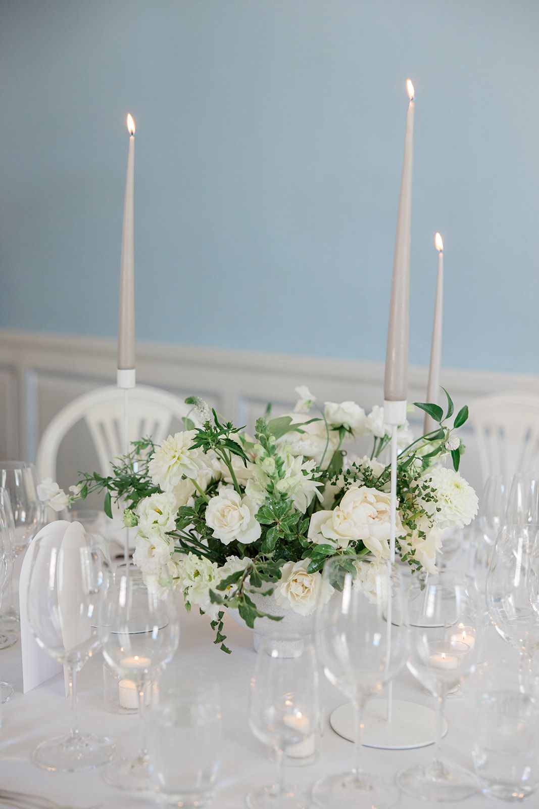 Elegant white table setting with tall taper candles and white floral centerpieces in glass vases on a minimalist tablescape.
