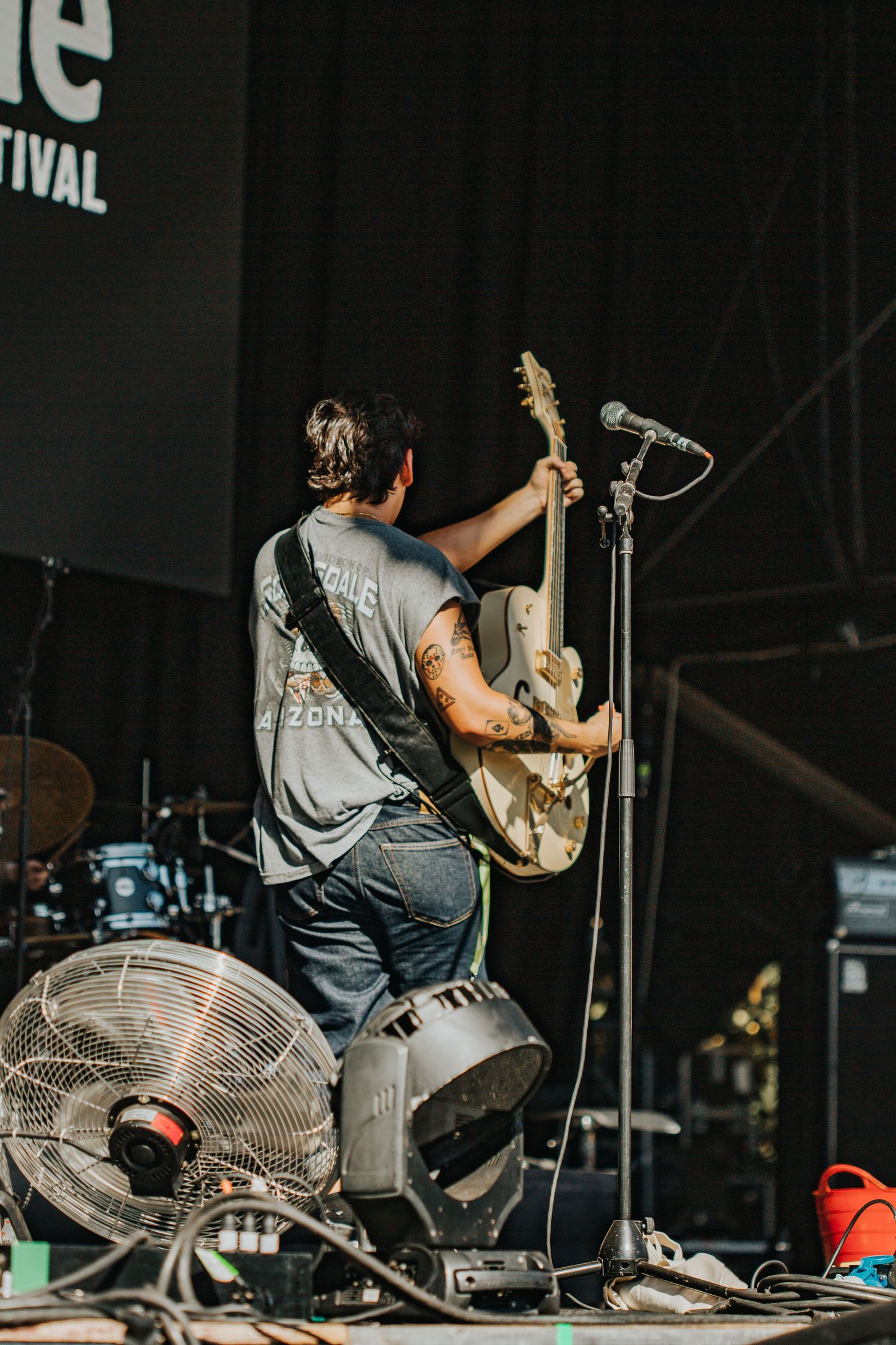 Tattooed guitarist seen from behind holding guitar up toward microphone on festival stage.