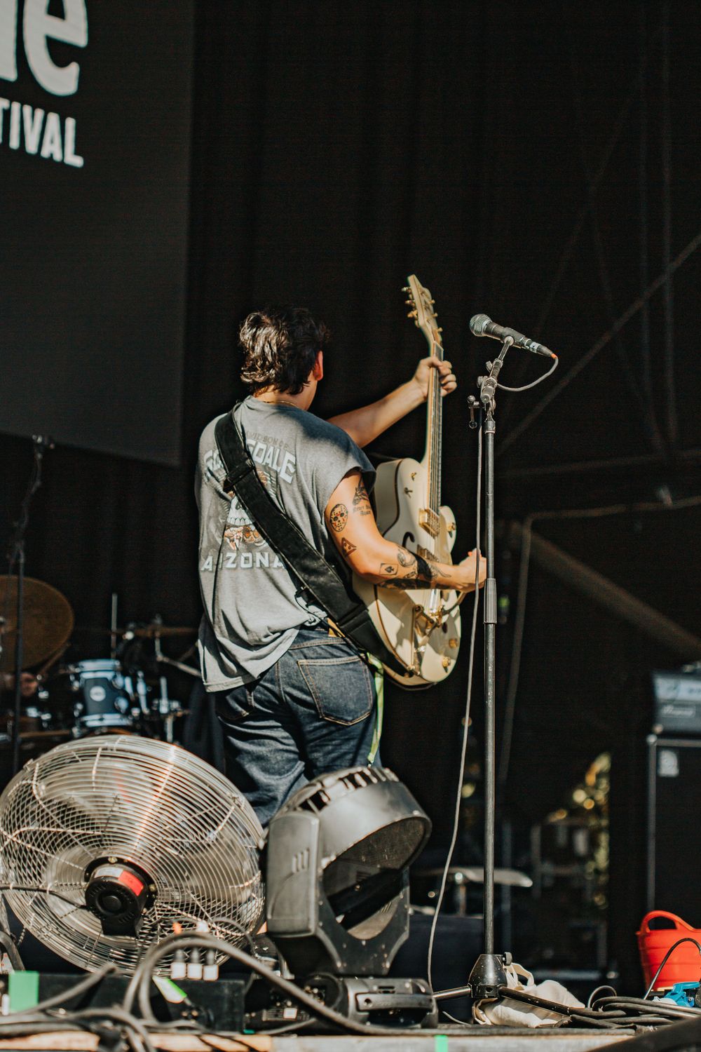 Tattooed guitarist seen from behind holding guitar up toward microphone on festival stage.