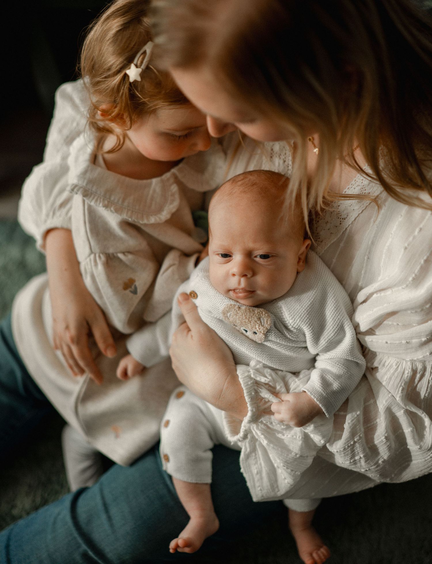 A mother in a white lace dress holds a newborn baby while a toddler girl in white looks on tenderly.