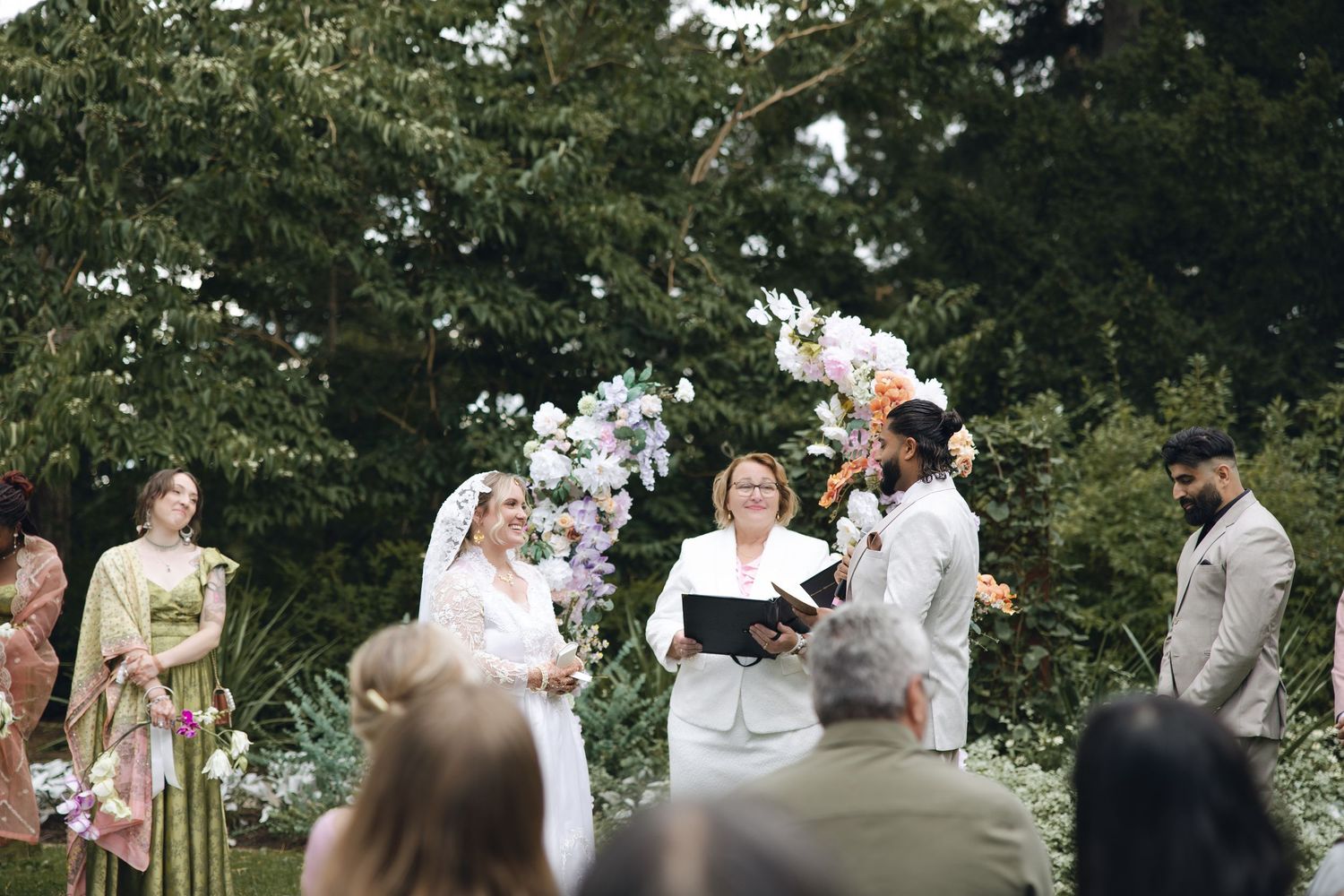 Wedding party poses together on lush green lawn with tall pine trees in background during summer celebration.