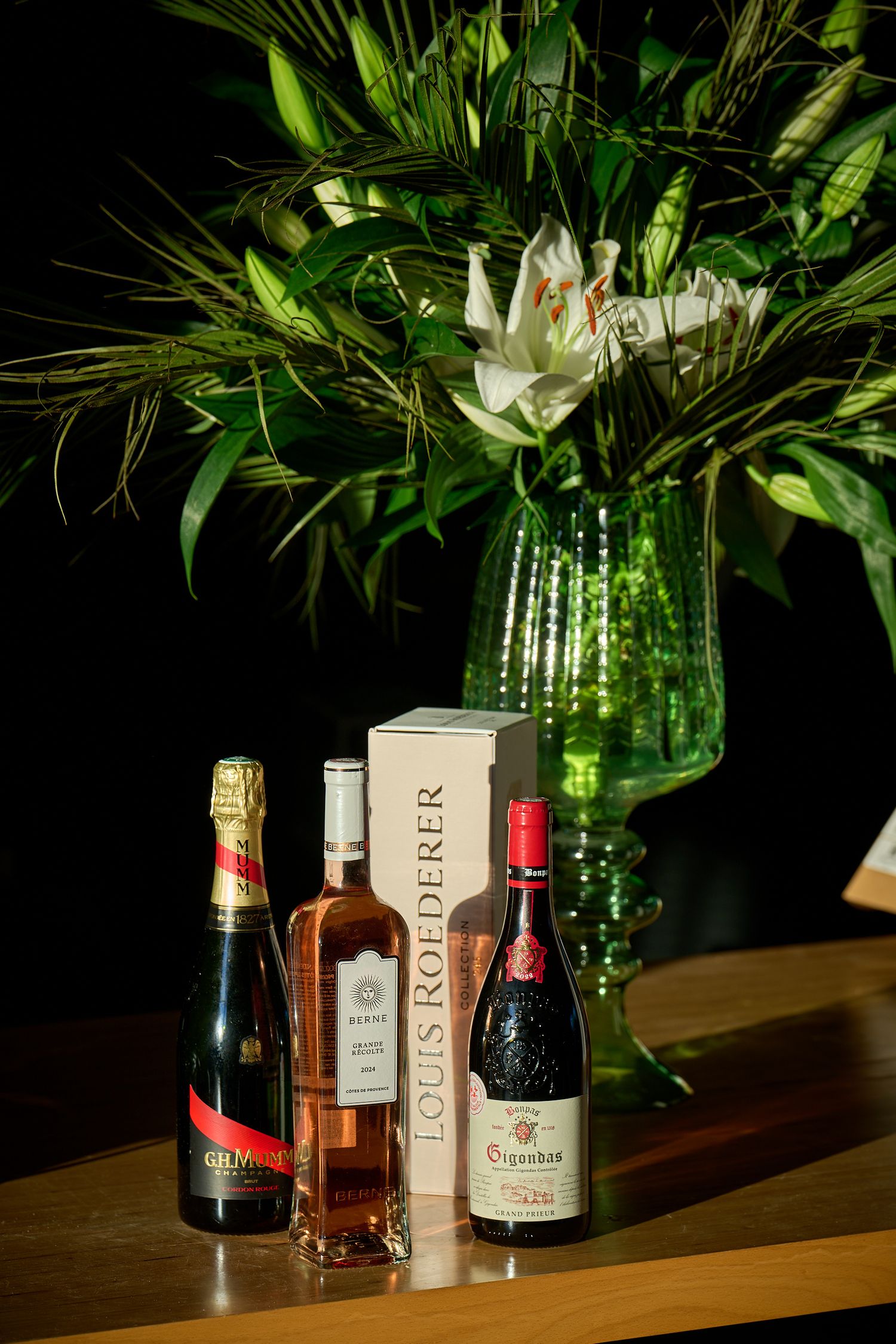 Elegant floral arrangement in green glass vase with wine and liquor bottles displayed on wooden table against dark background.