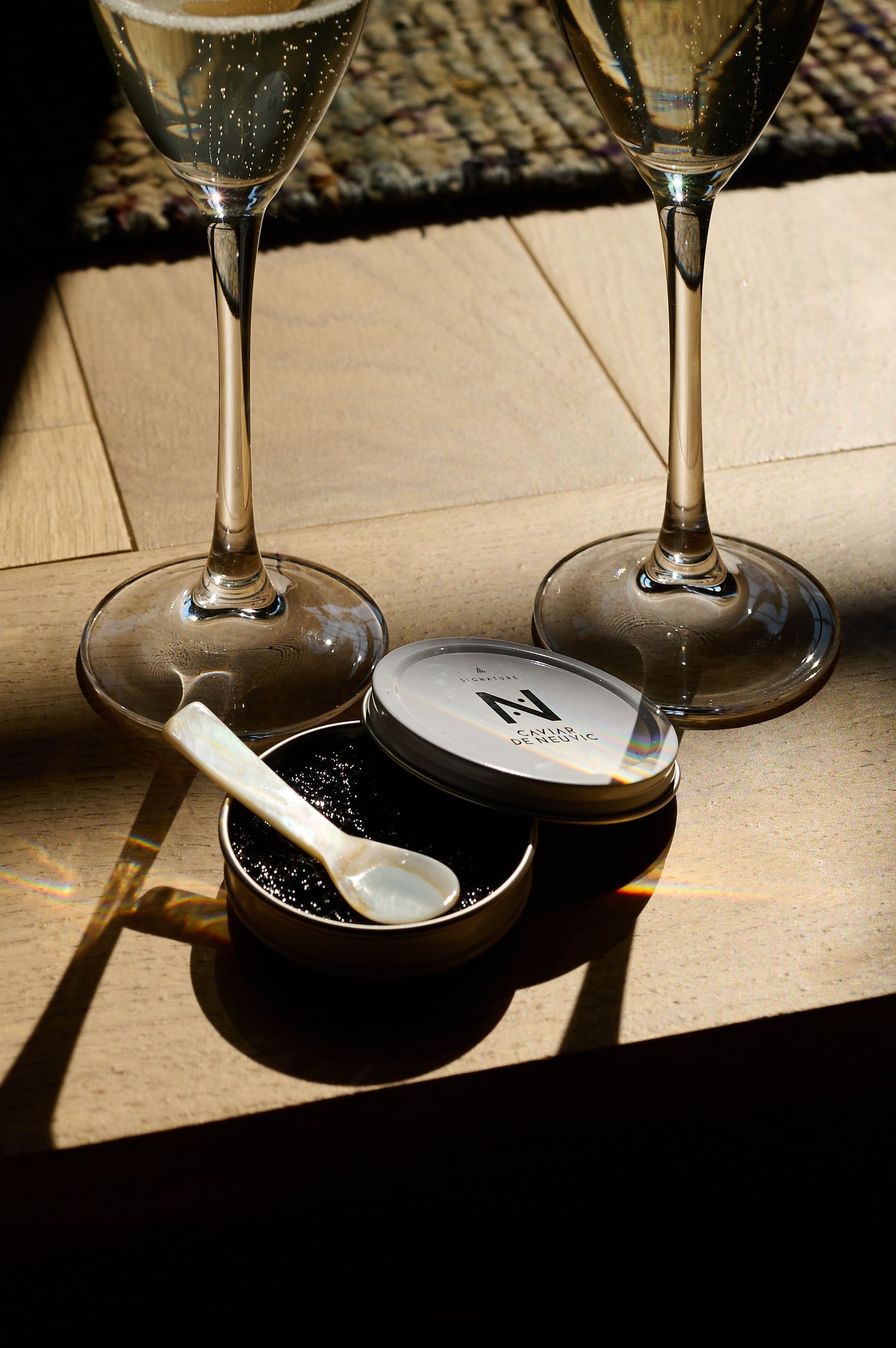 Two champagne glasses cast shadows beside a black caviar tin with a small spoon on a textured surface.