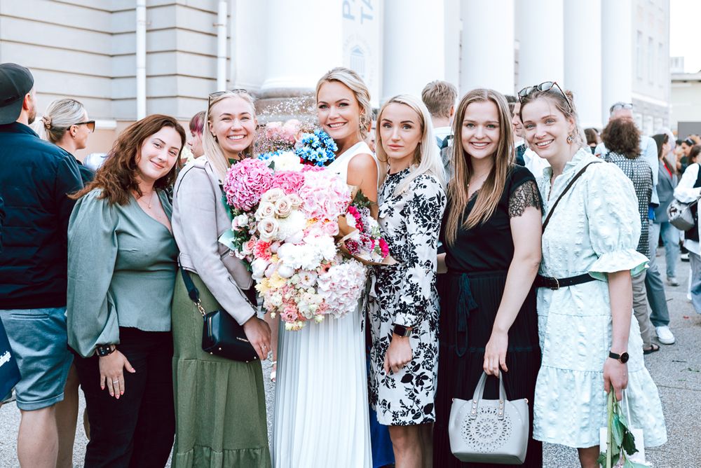 A group of wedding guests in floral dresses and formal attire gather together for photos outside a white building.