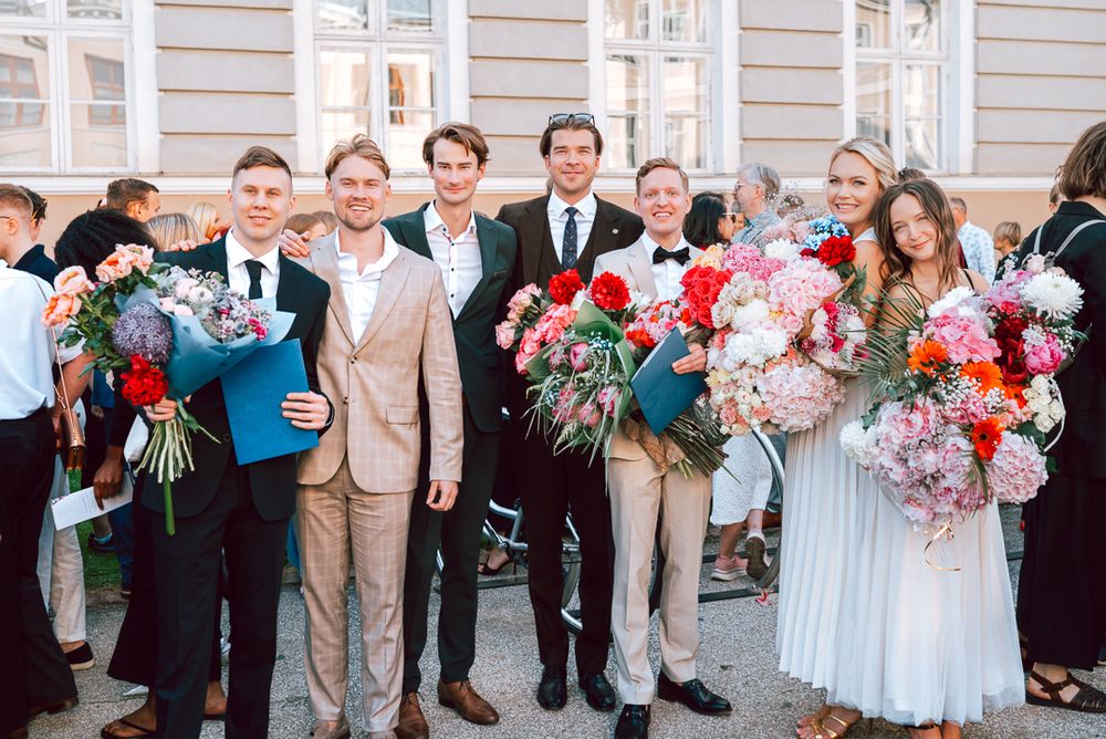 Wedding party members in formal attire hold vibrant bouquets of pink and white flowers outside an elegant building.