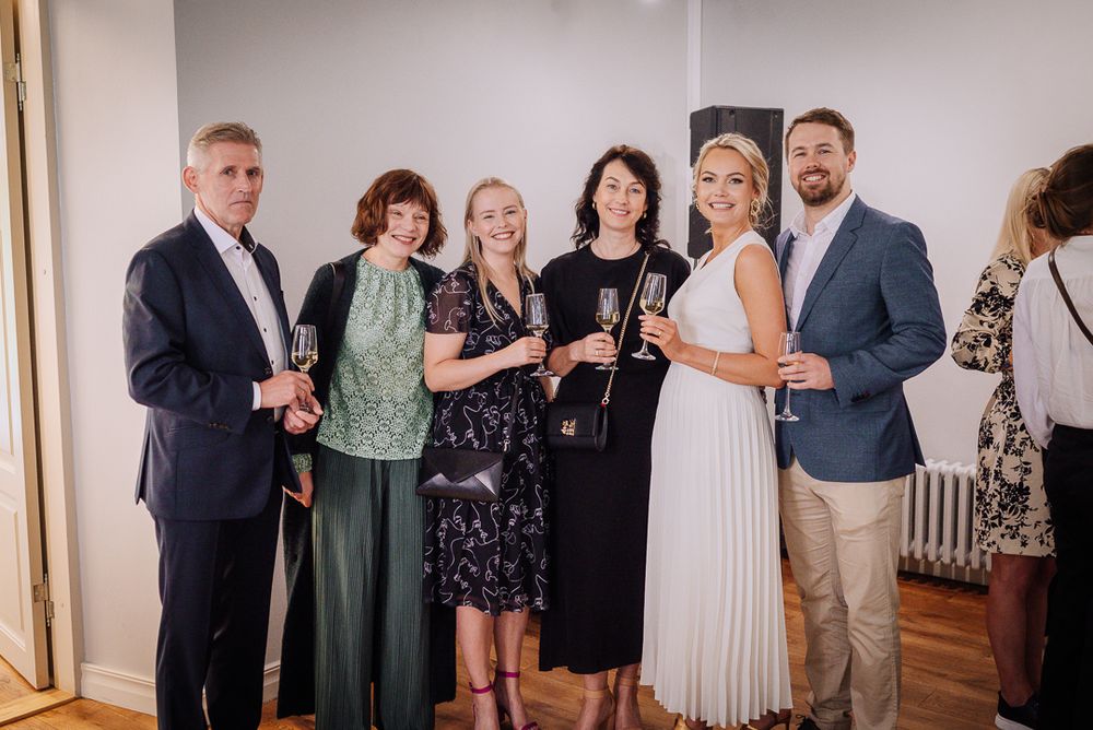 Wedding guests in formal attire stand together in a line against a white wall during a celebration.