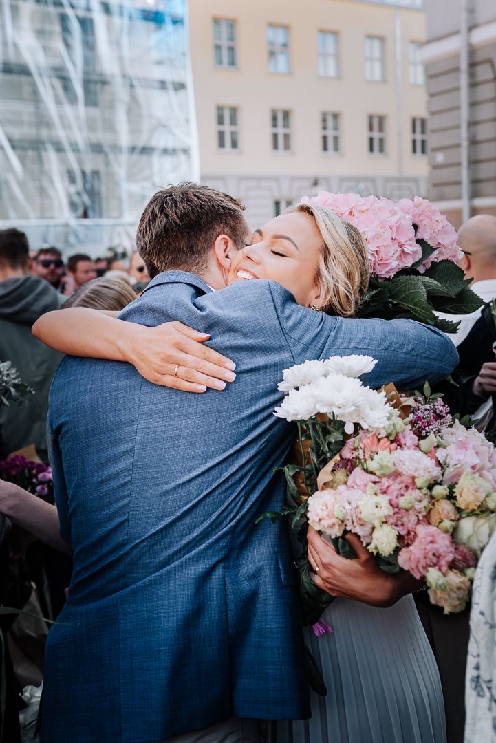 Wedding guests share emotional embraces and hugs while holding pink and white flower bouquets at an outdoor celebration.