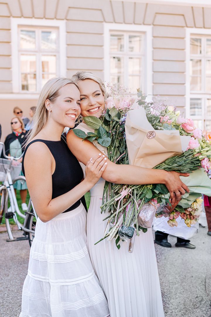 Two people in white dresses embrace while holding a large bouquet of pink and white flowers outdoors.