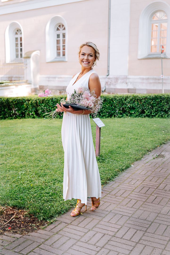 A person in a flowing white pleated maxi dress stands on a paved path near a classical building with arched windows.