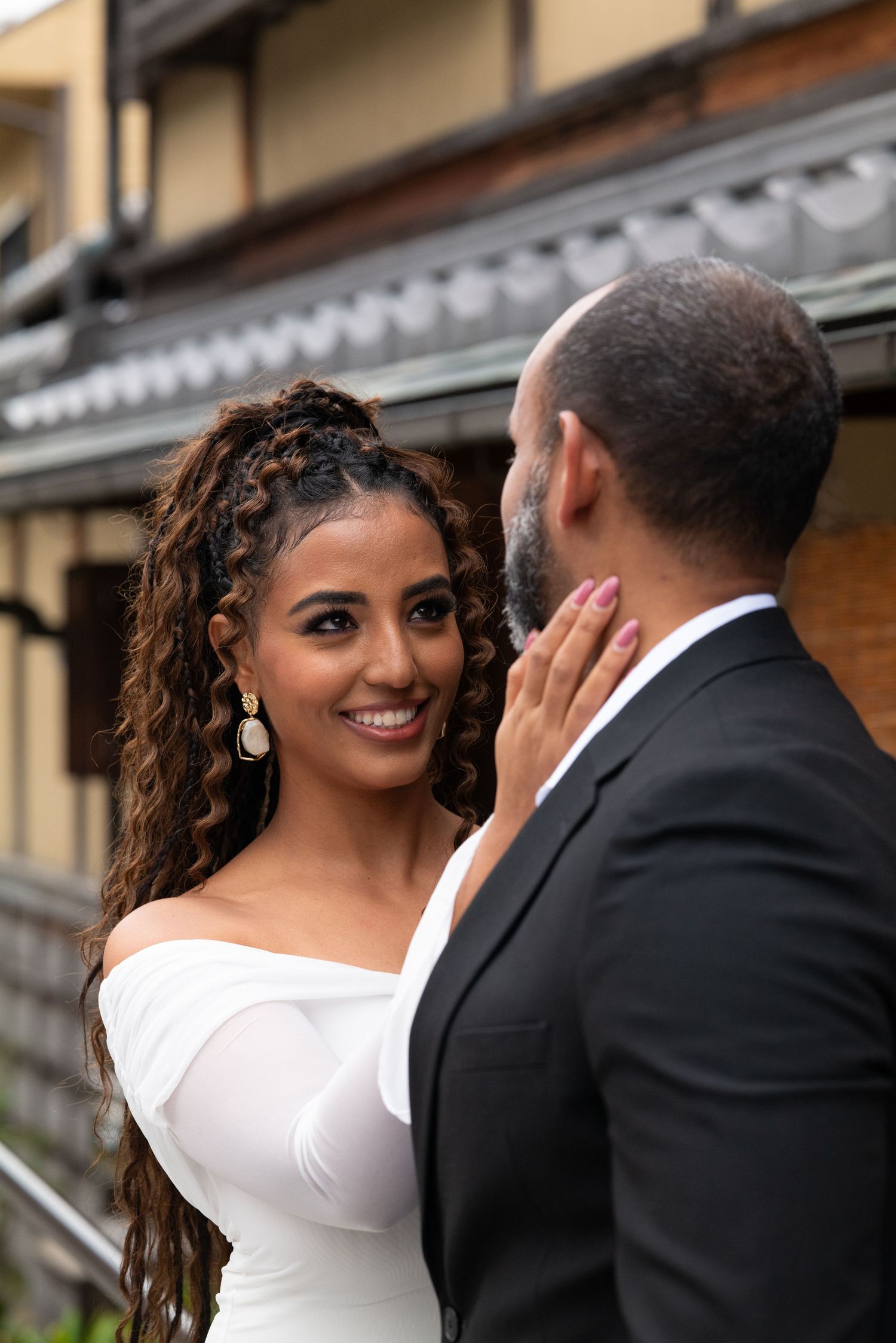 Smiling bride in off-shoulder white dress touches grooms face tenderly outside a traditional Japanese building.