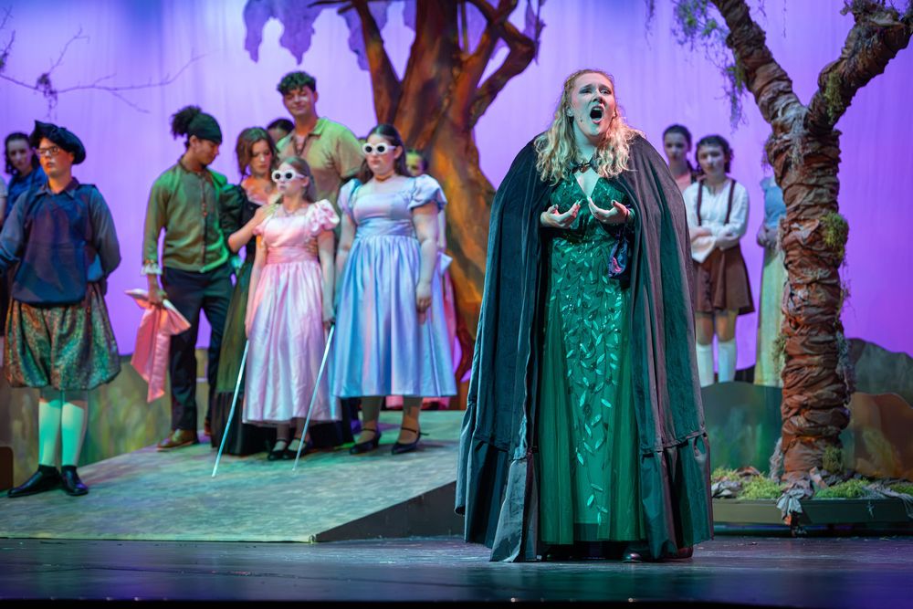 Stage performers in medieval-style costumes during a theatrical production with purple lighting.