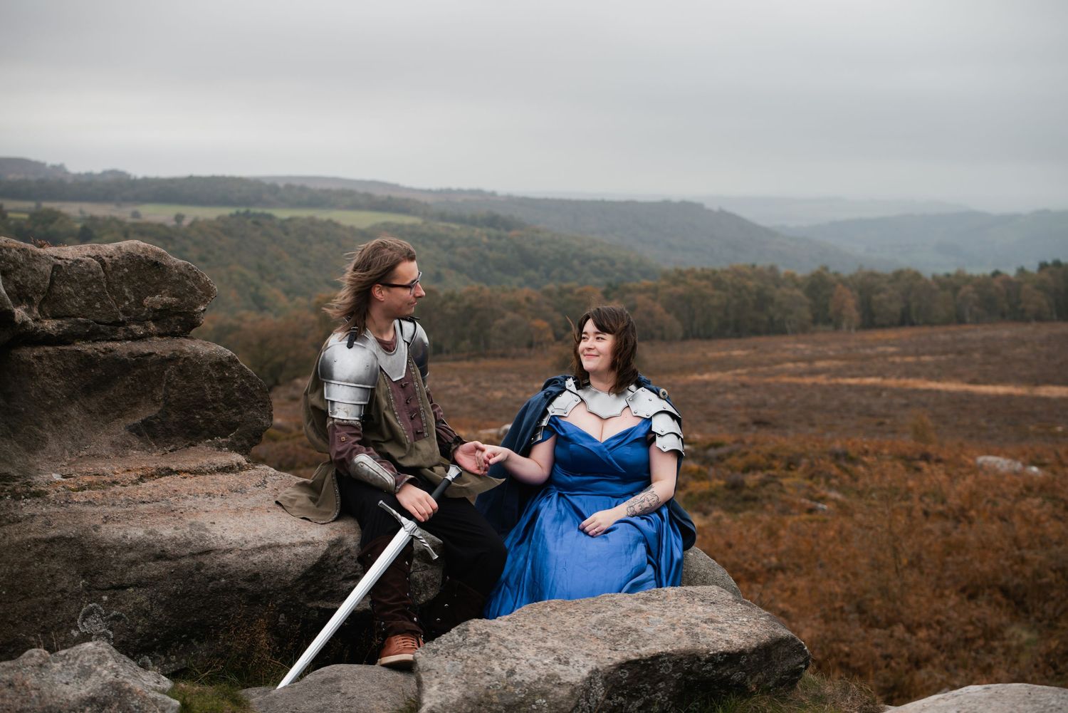 A couple in fantasy wedding attire poses on rocky terrain with mountains in the background.
