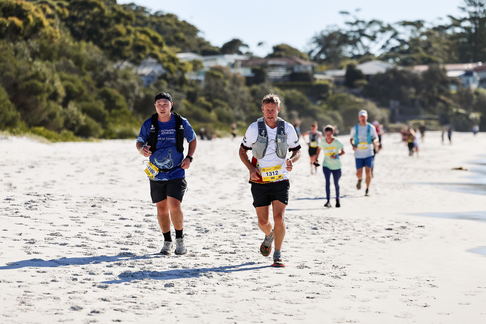 Runners competing in a beach race on white sand with houses and trees visible in the background.