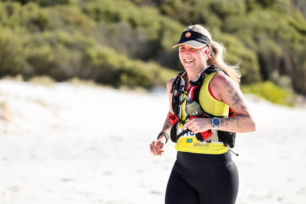 Smiling female trail runner in yellow vest and black leggings jogging on sandy path with trees in background.