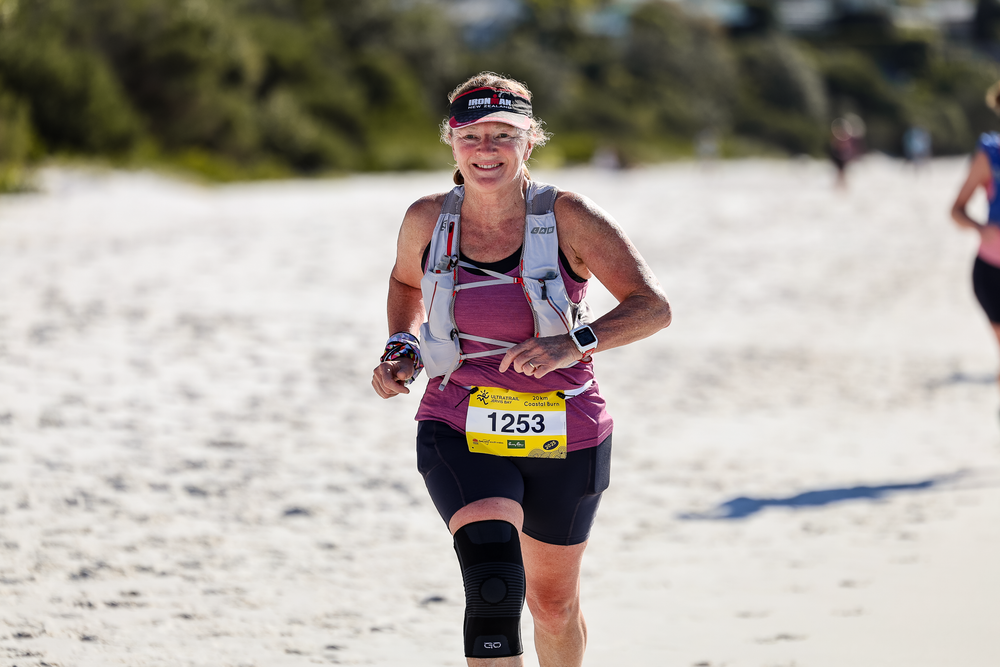 Female runner #1253 competing in a beach triathlon race, wearing a pink top and knee brace on a sunny day.
