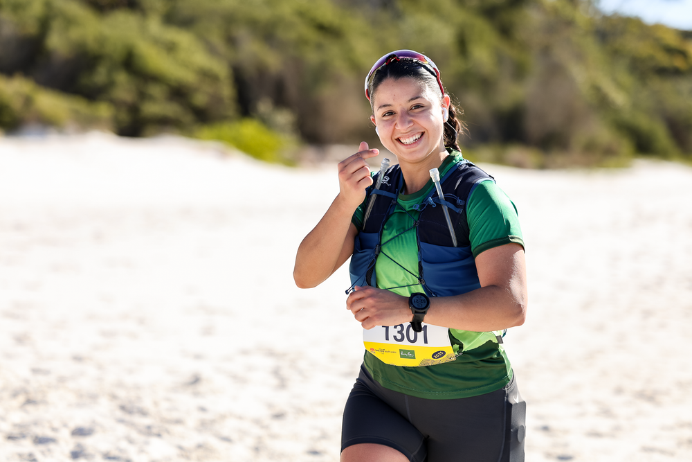 Smiling female trail runner wearing bib 1301 and green shirt running on sandy beach during race.
