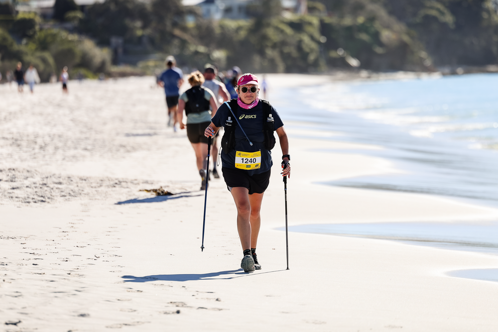 Athlete using trekking poles runs on a sunny beach during a race, wearing a yellow bib number and black outfit.