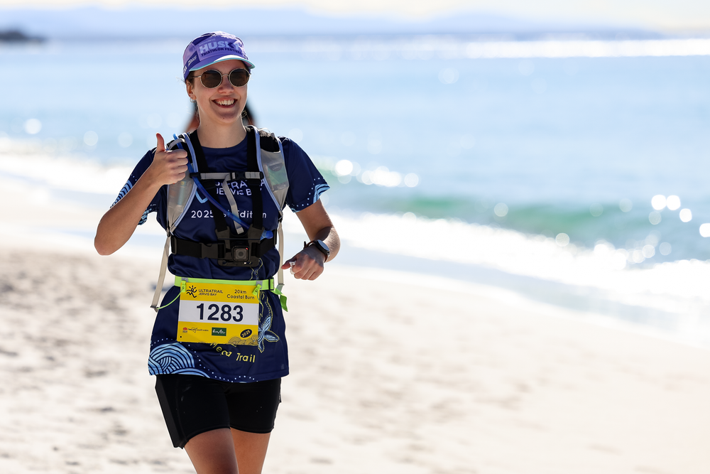 Female runner #1283 smiling and giving thumbs up on sandy beach during a marathon race.
