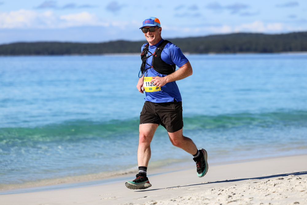 Man running a beach marathon race wearing blue shirt, race bib, and cap on sunny white sand shore.