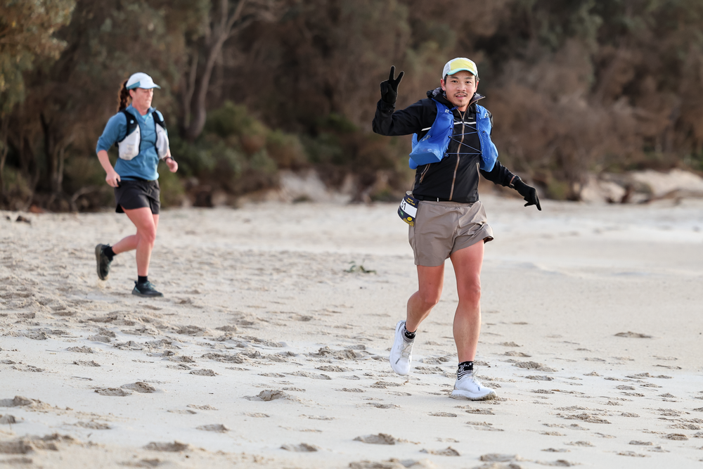 Two women running on a sandy beach wearing hydration vests and athletic gear during an outdoor trail race.