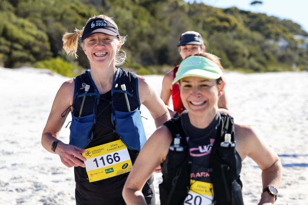 Two women runners wearing race bibs 1166 and 20 smile during a trail race on a sandy beach path.