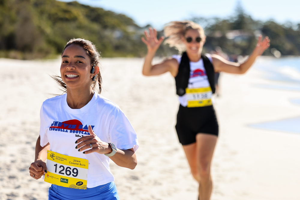 Two women running on a sandy beach in a race, wearing numbered bibs, smiling and celebrating outdoors.