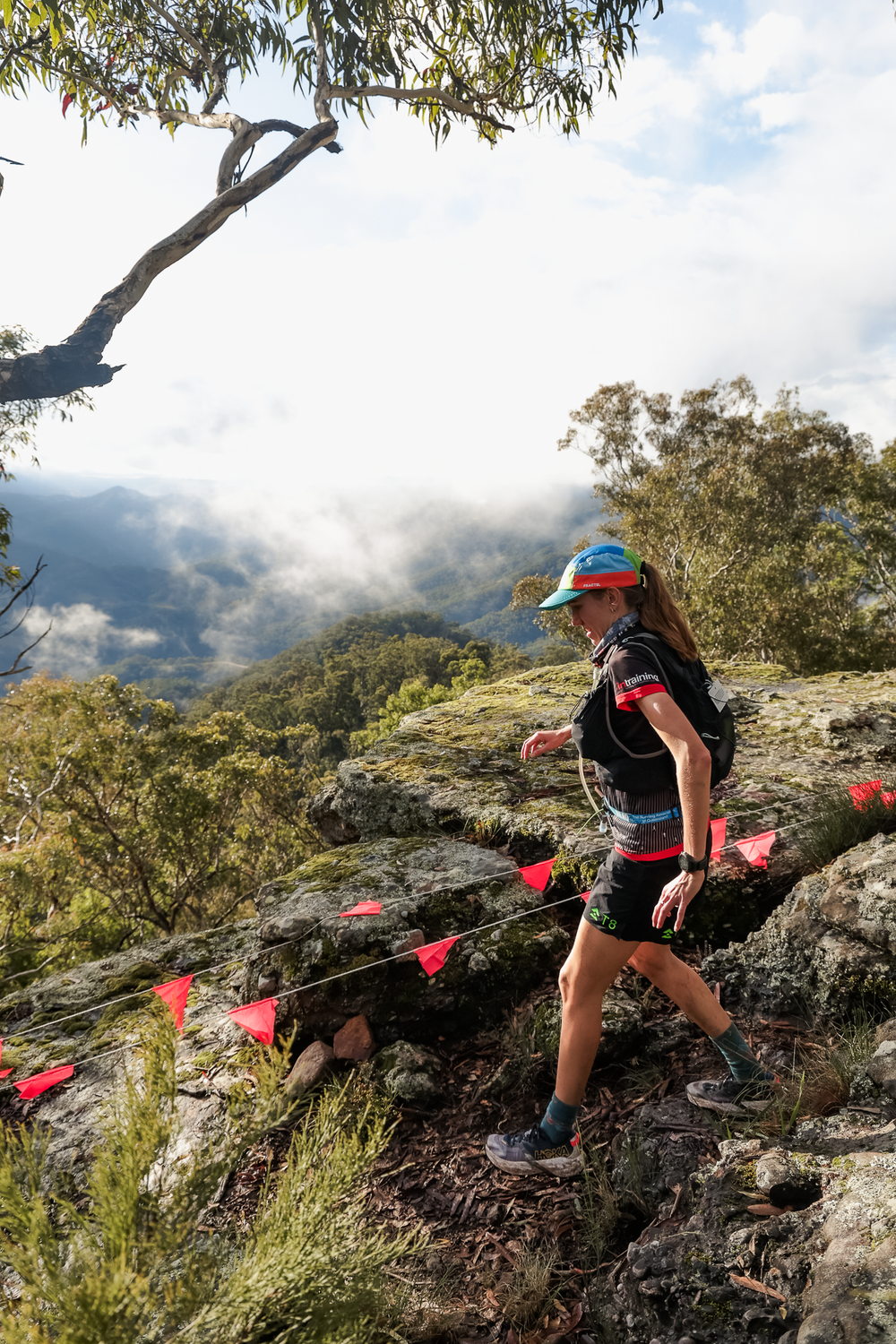 Female trail runner navigating rocky mountain path with red flags and misty valley views.