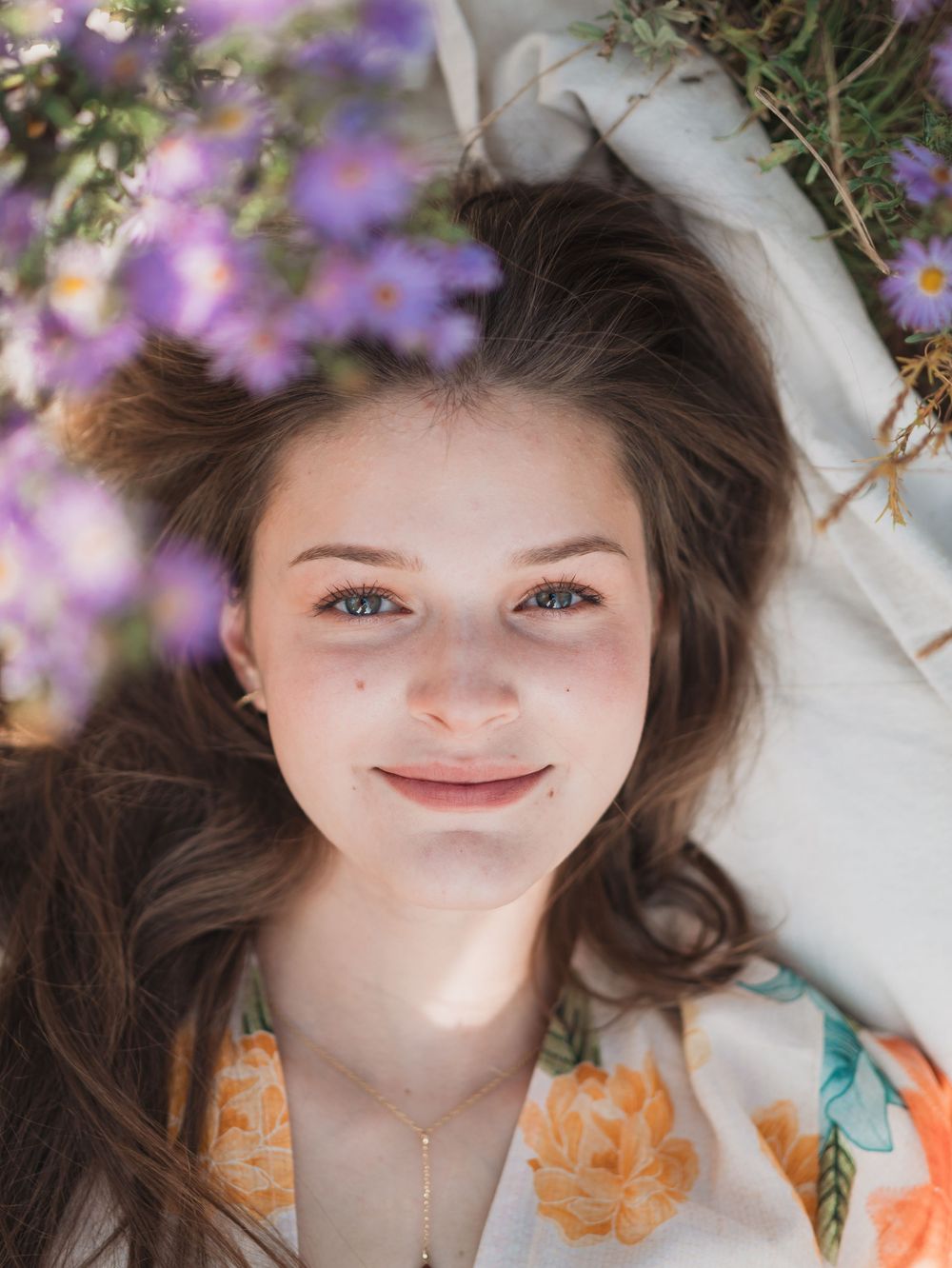 A smiling figure lying on white fabric with purple flowers in the background, wearing a floral print top.