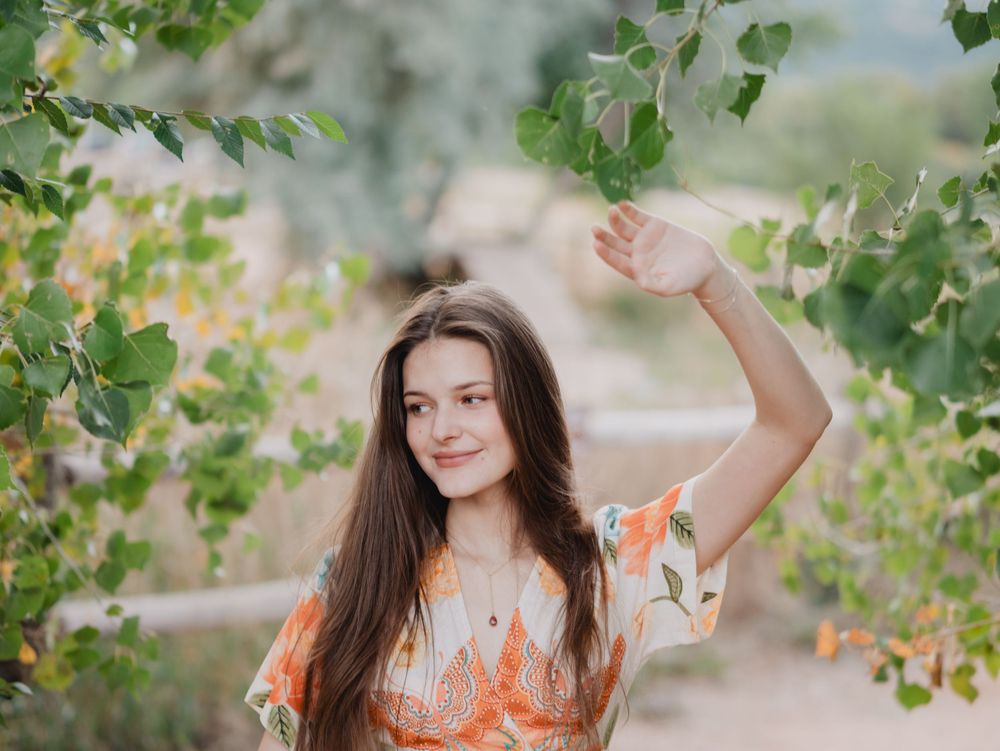 A woman in an orange dress poses in a natural outdoor setting surrounded by green foliage and sunlit trees.
