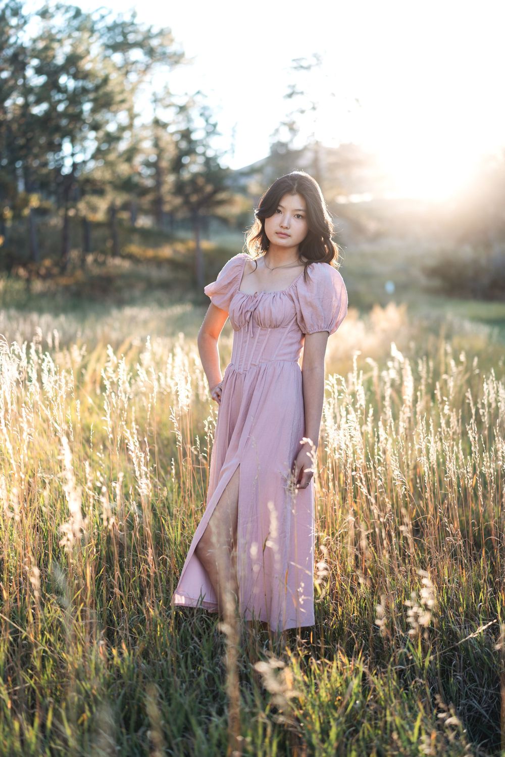 A person in a pink dress stands among tall grass in a golden-lit meadow at sunset.