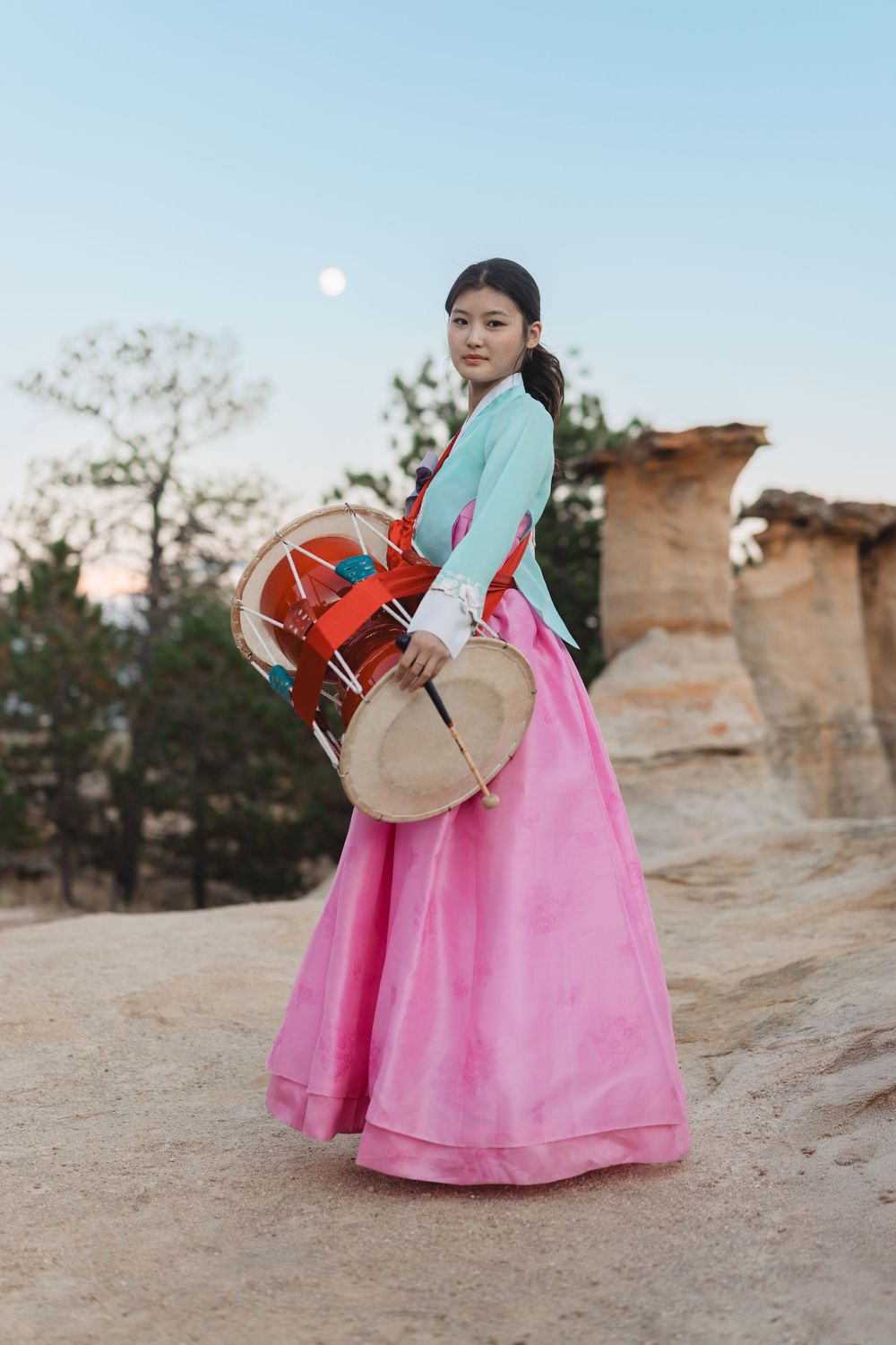 A person in a bright pink hanbok dress carries traditional Korean drums against a desert rock formation backdrop.