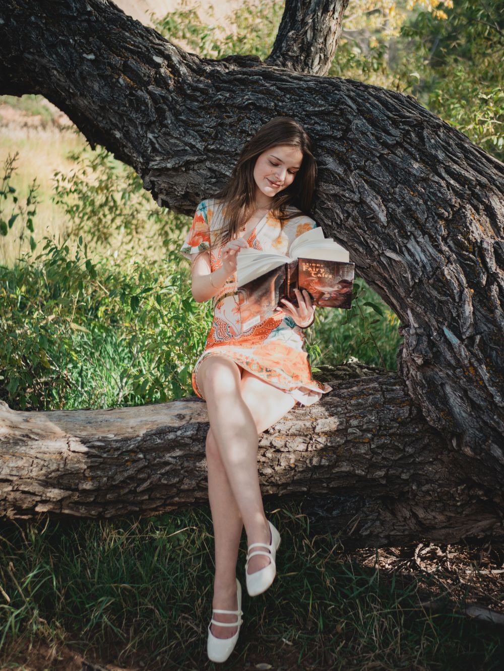 A person in a peach-colored dress sits on a fallen tree trunk in a sunlit outdoor setting.
