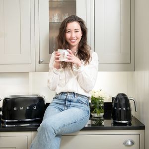 Woman in white sweater and jeans relaxes on kitchen counter with coffee and casual poses in modern kitchen.
