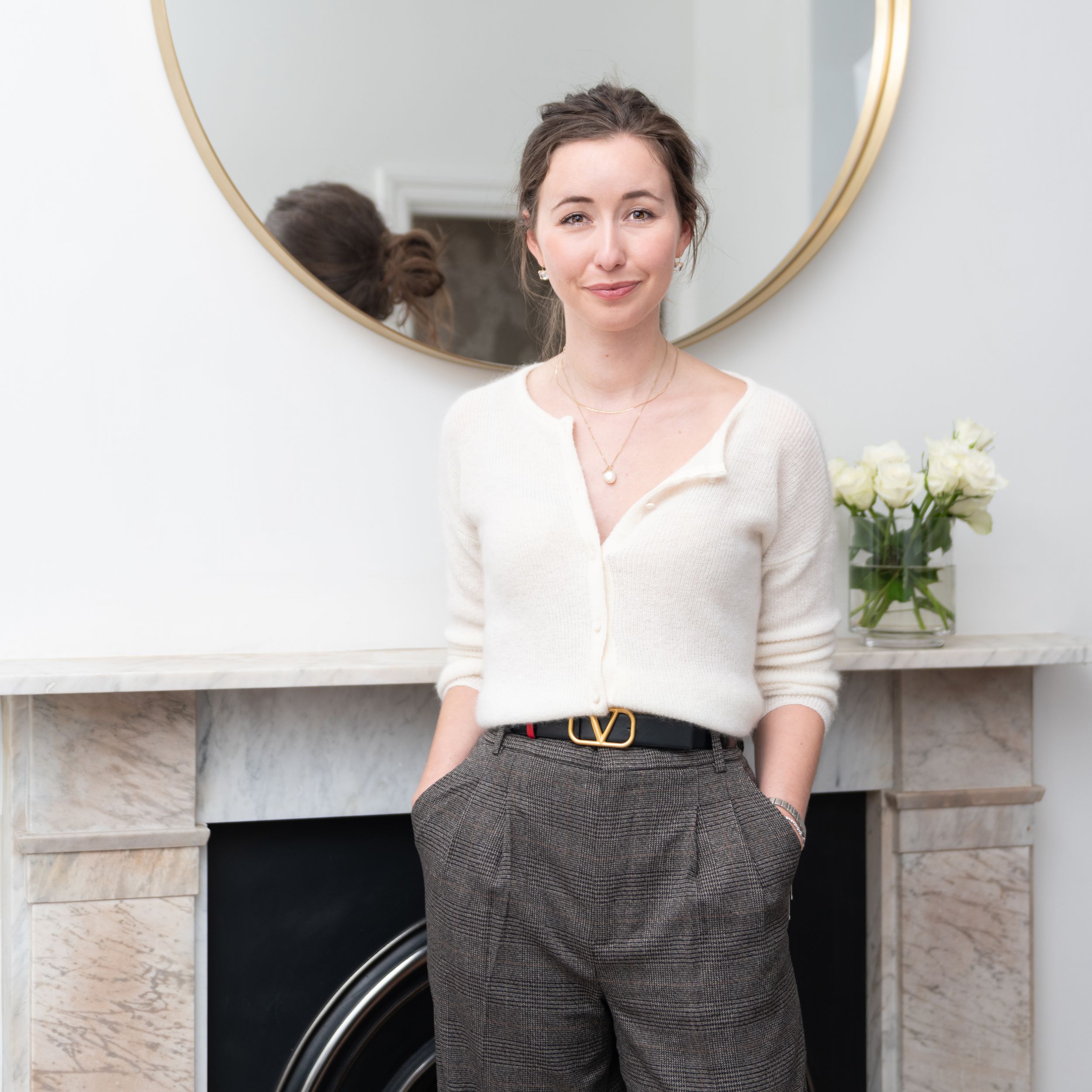 A person in a white cardigan and gray trousers stands casually by a fireplace with a round mirror and white roses.