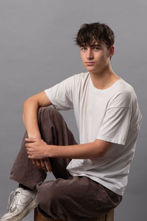Young person in white t-shirt and brown pants poses against gray background in artistic portrait series.