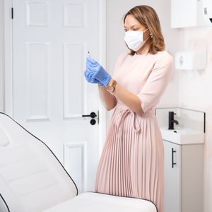 Healthcare worker in protective mask and gloves wearing pink dress prepares for medical procedure in examination room.