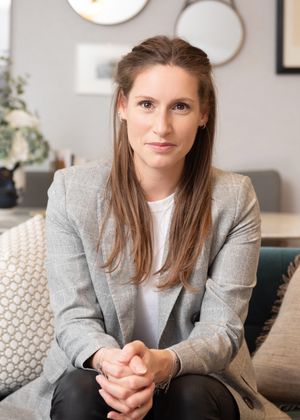Professional portrait of someone in a gray blazer and white shirt sitting on a couch in a modern living room setting.