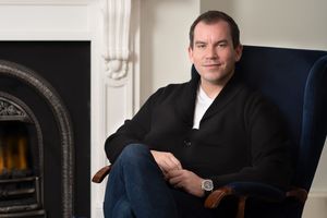 Person in a black sweater sits comfortably in a blue chair next to a fireplace in a bright room.