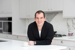Someone in a black sweater leans casually on a white marble kitchen counter with modern cabinets.
