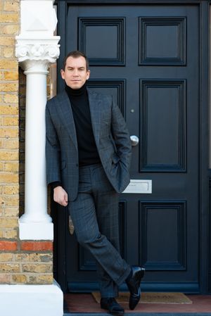 Person in dark suit stands beside black paneled door and yellow brick facade with white columns.