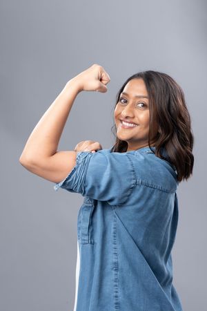 A person in a denim shirt flexes their bicep while smiling over their shoulder against a gray backdrop.