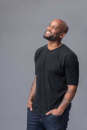 A person wearing a black t-shirt and jeans smiles joyfully against a gray background.