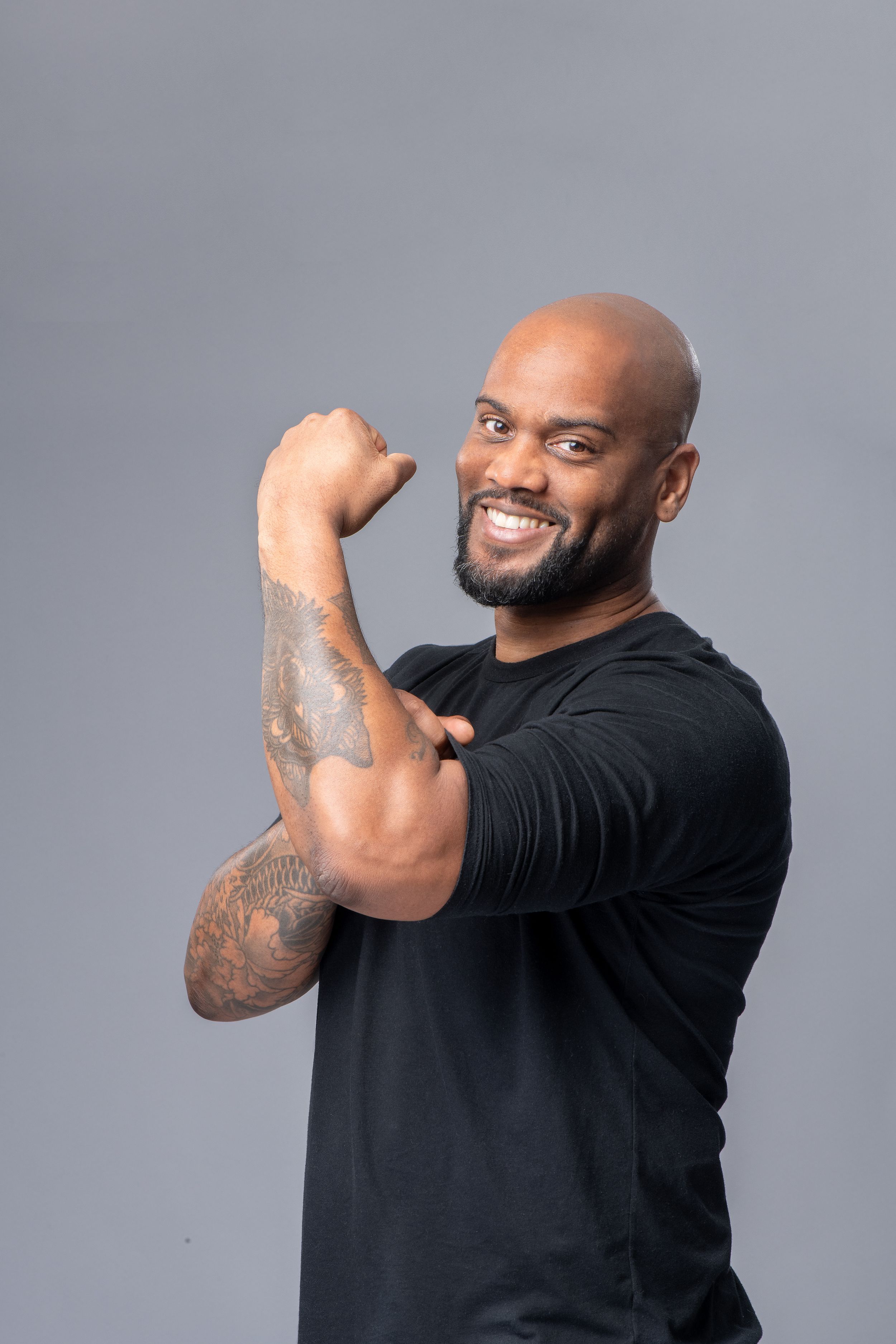 Man in black shirt demonstrates arm flexing poses while smiling against gray backdrop.