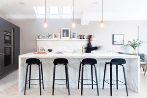 Modern white kitchen with marble island counter featuring four black bar stools and pendant lighting.